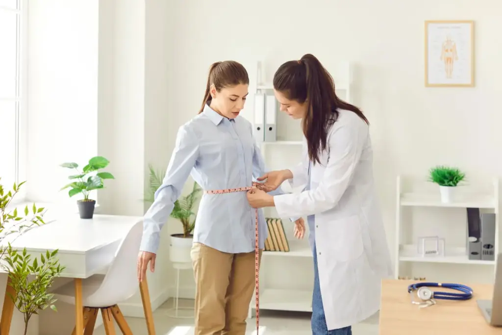 Healthcare professional measuring a young woman’s waist during a wellness or weight management consultation in a medical office.
